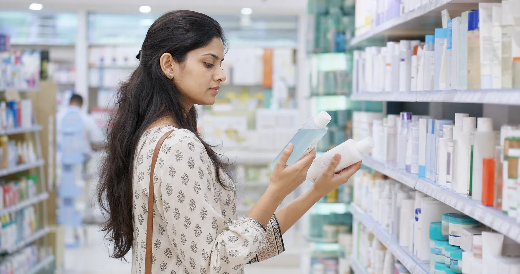 Indian shopper thoughtfully comparing two unbranded beauty products in a pharmacy aisle