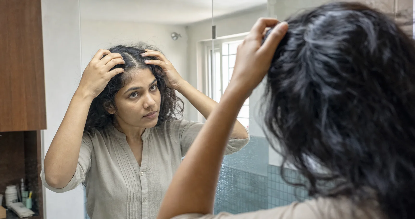Indian woman gently parting her natural hair while inspecting her scalp in a bathroom mirror